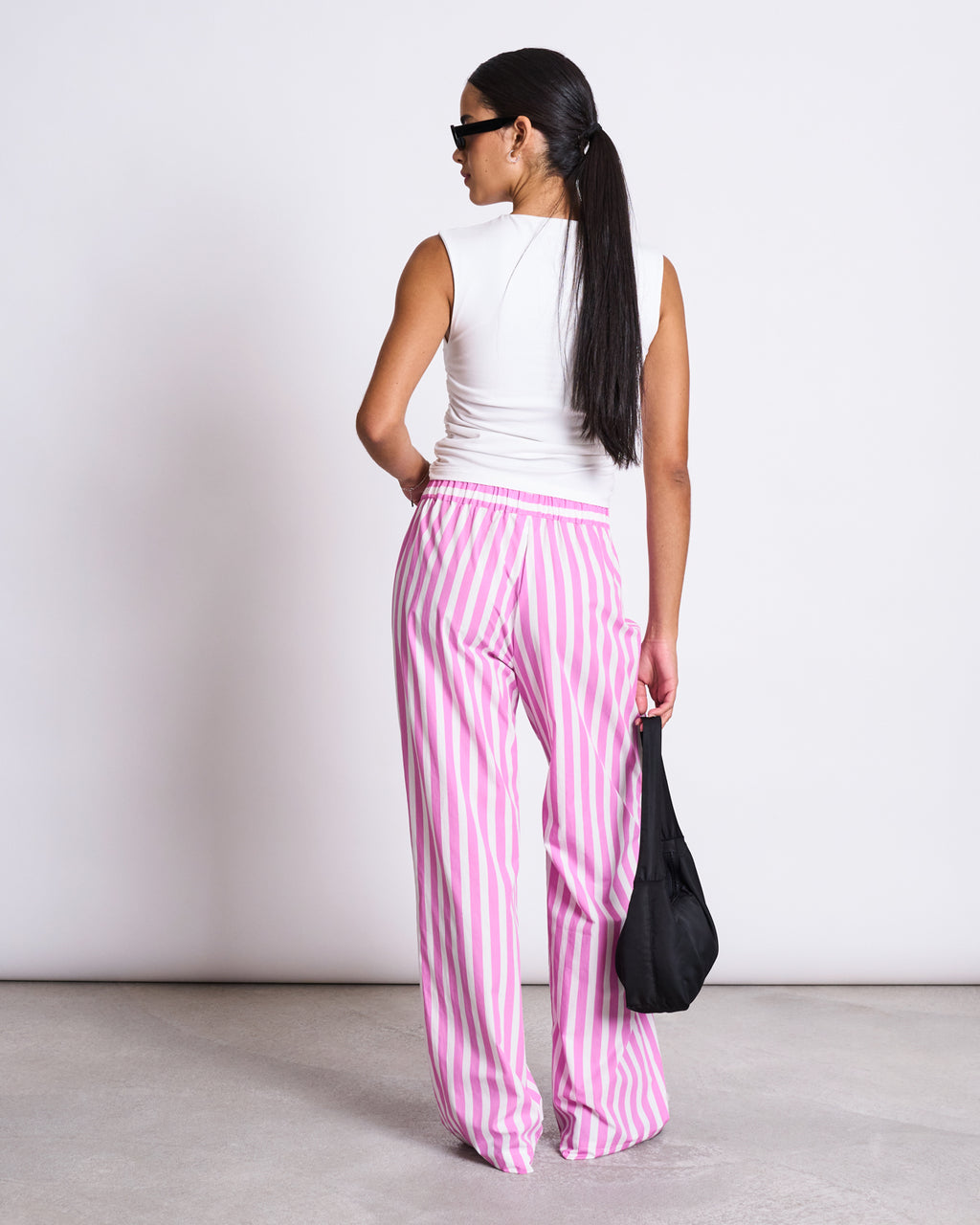 A woman with long dark hair, sunglasses, and a white sleeveless top stands on a light floor against a white wall. She is wearing jan-n-junes COTTON PANTS SOBY WHITE HOT PINK STRIPED GOTS and holding a black bag.