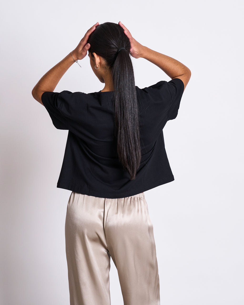 A woman with long dark hair in a ponytail, facing away with hands on her head, wears an oversized jan-n-june BOXY T-SHIRT TEMEL BLACK and light beige satin pants against a plain white background.