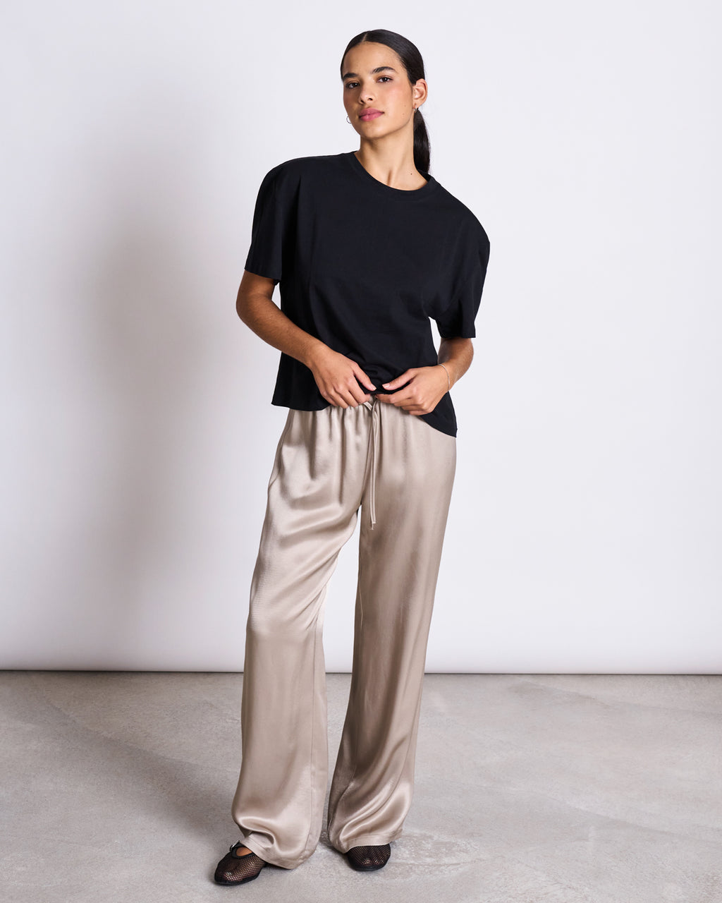 A woman stands against a white wall wearing the jan-n-june BOXY T-SHIRT TEMEL BLACK, paired with beige satin wide-leg pants and black flats. She has dark hair tied back and looks ahead with a neutral expression.