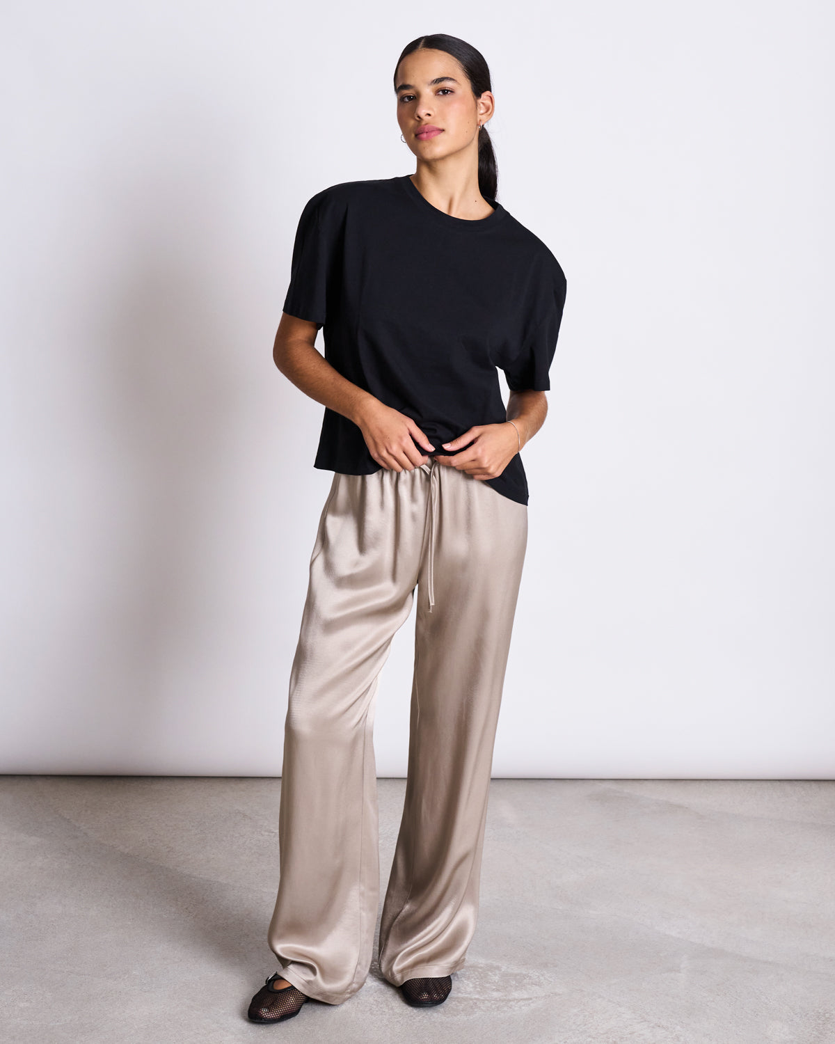 A woman stands against a white wall wearing the jan-n-june BOXY T-SHIRT TEMEL BLACK, paired with beige satin wide-leg pants and black flats. She has dark hair tied back and looks ahead with a neutral expression.