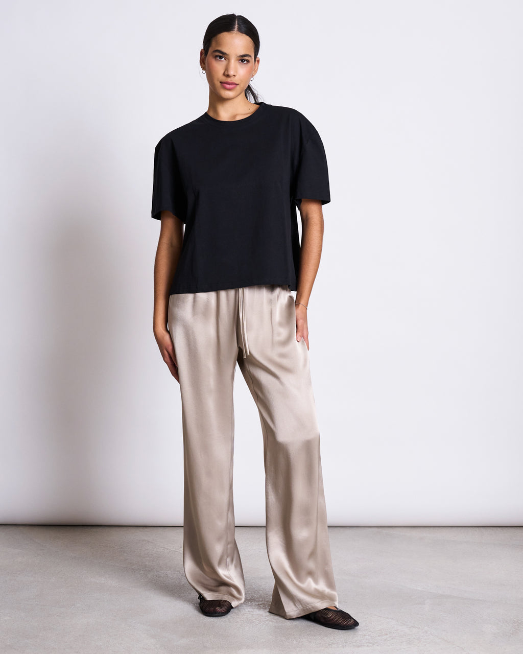 A woman stands against a white background wearing jan-n-junes BOXY T-SHIRT TEMEL BLACK, beige satin wide-leg pants, and dark flat shoes, looking at the camera with a relaxed posture.