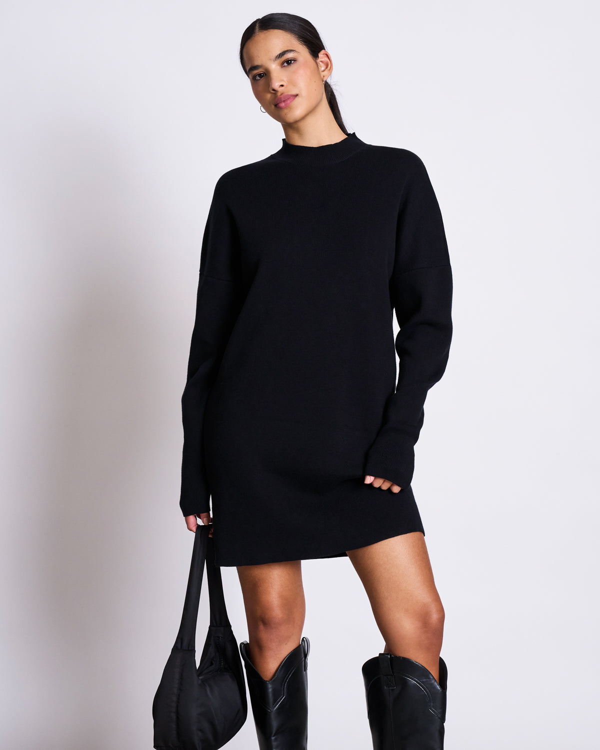 A woman with dark hair pulled back poses confidently in the jan-n-june KNIT DRESS NUNA BLACK GOTS, a long-sleeve oversized black organic cotton dress, paired with knee-high black boots and a matching black bag against a plain background.