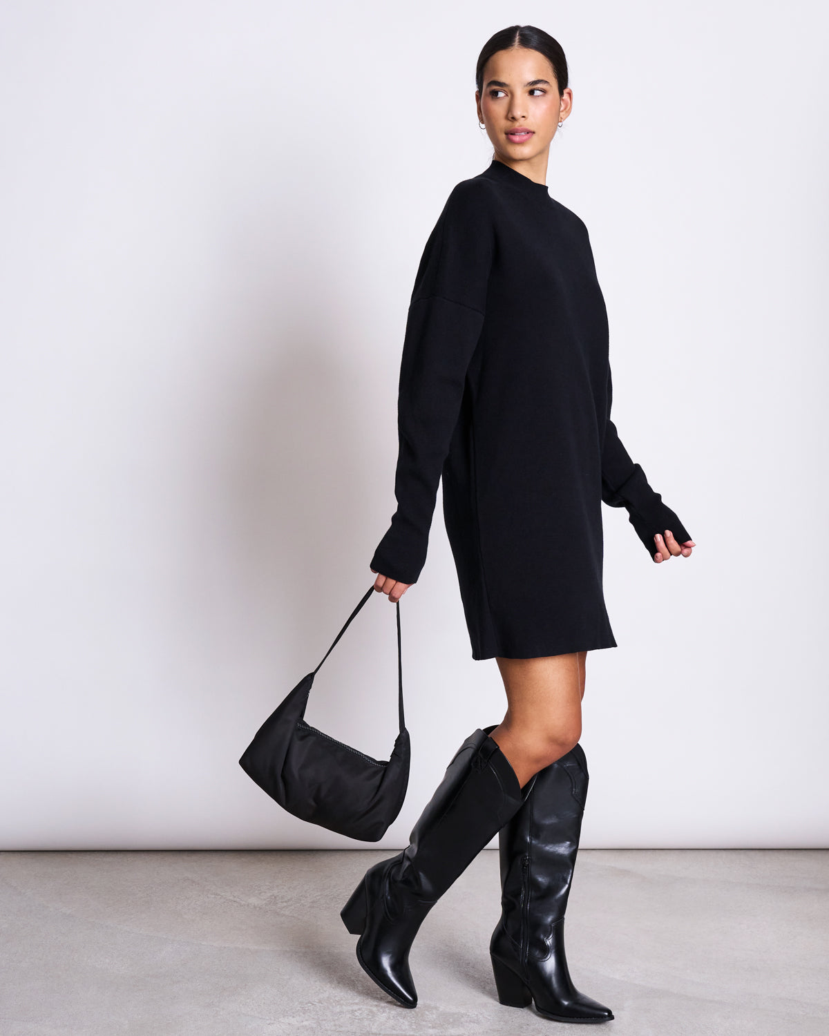 A woman in the jan-n-june KNIT DRESS NUNA BLACK GOTS, an organic cotton long-sleeve oversized dress, with black knee-high boots and a black handbag, stands against a white background, looking over her shoulder.