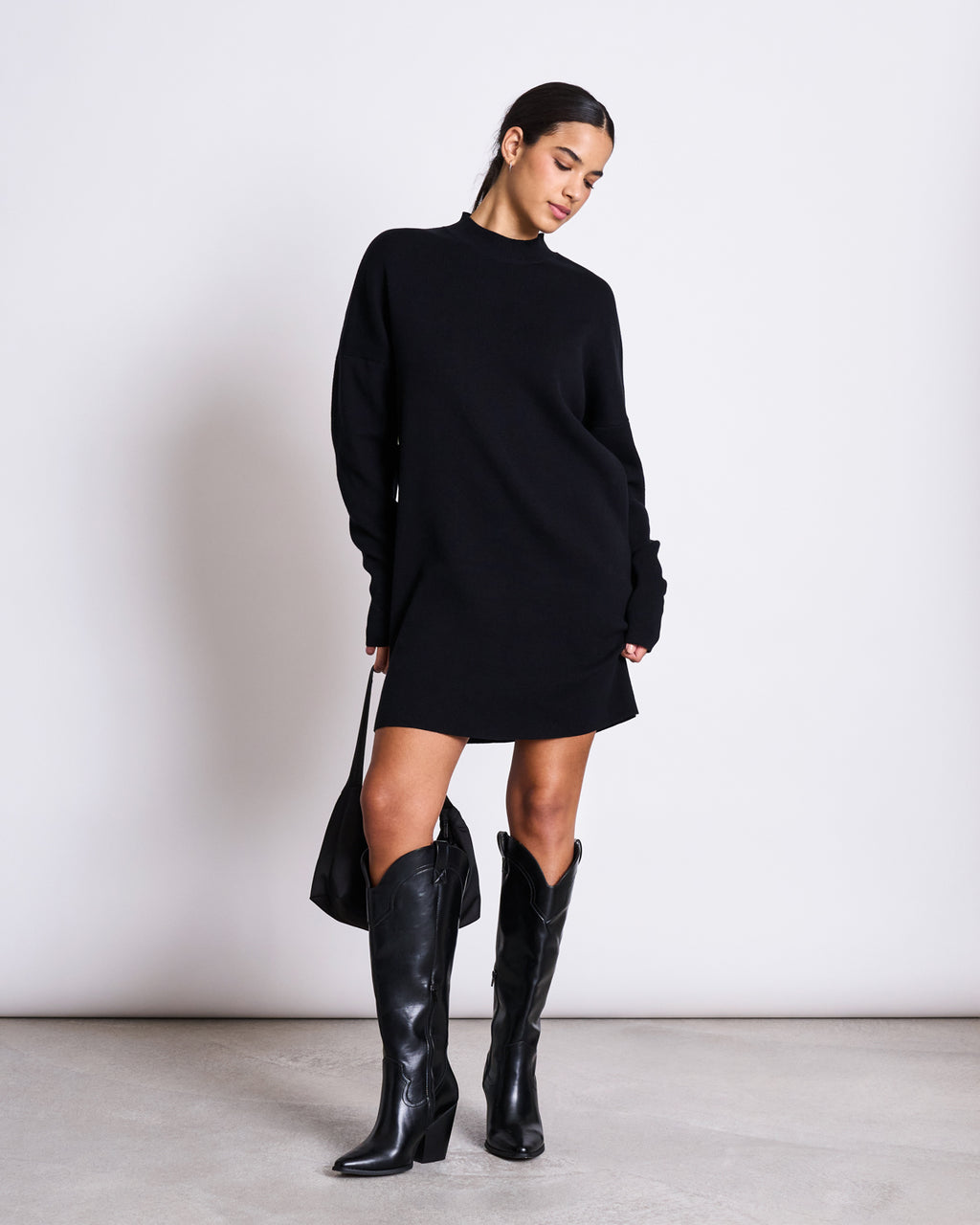 A woman stands against a white background, wearing the jan-n-june KNIT DRESS NUNA BLACK GOTS, paired with black knee-high cowboy boots and holding a black handbag. She looks down with a relaxed expression.