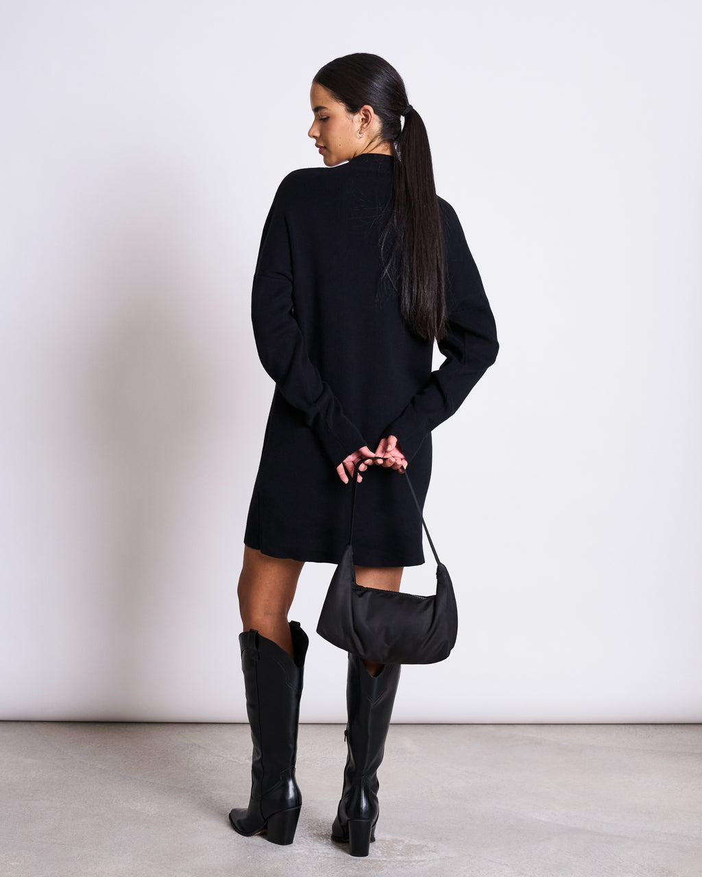 A woman with long dark hair in a ponytail, facing away, wears the jan-n-june KNIT DRESS NUNA BLACK GOTS with black knee-high boots and holds a small black purse behind her back against a plain light background.