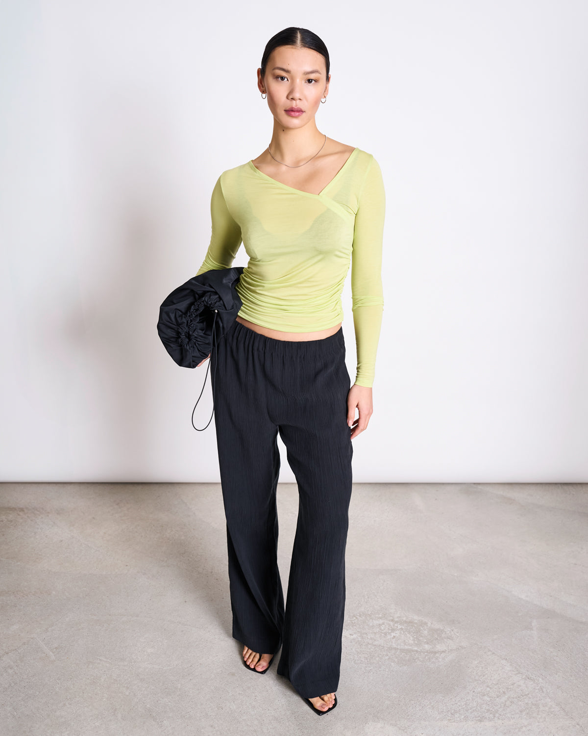 A woman stands in a minimal studio wearing the jan-n-june ASYMMETRICAL LONGSLEEVE CASMA GRANNY SMITH, paired with loose black pants and sandals. She holds a black bag, her hair pulled back and expression neutral.