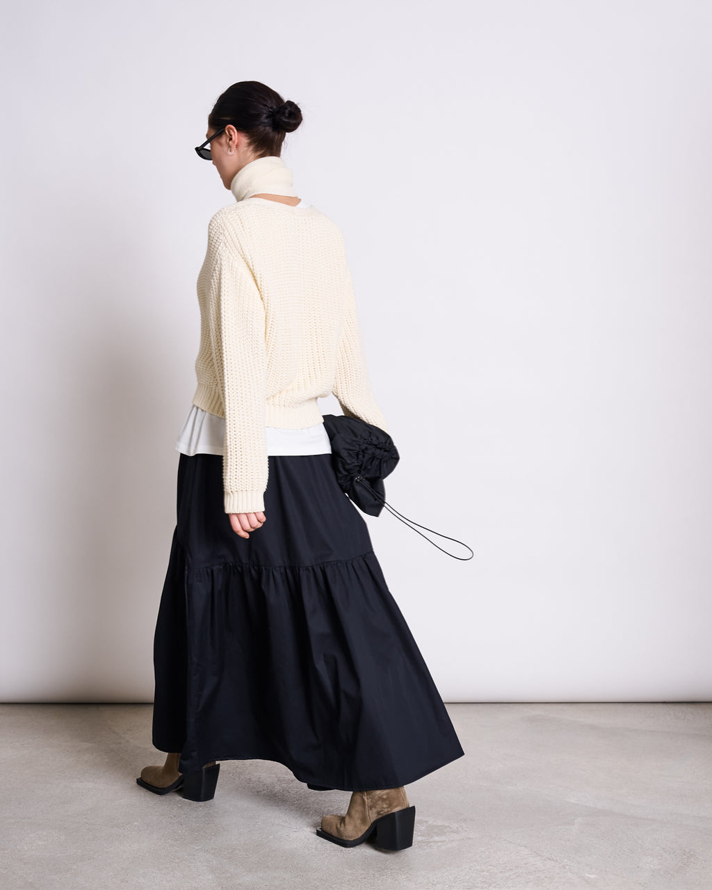 A person with dark hair in a bun, glasses, and a jan-n-june CHUNKY CARDIGAN LENA OFFWHITE GOTS walks away from the camera in a minimalist indoor setting, paired with a long black skirt, beige boots, and holding a small black bag.