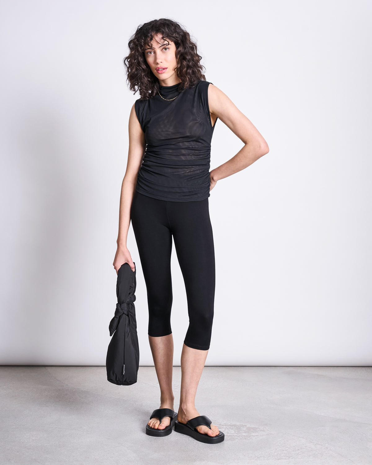 A woman with curly hair wears the jan-n-june RUFFLE MESH TOP LAGUNA BLACK, black cropped leggings, and sandals, standing on a gray floor and holding a folded black bag against a plain white background.