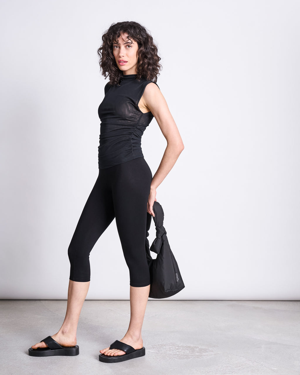 A woman with curly hair stands against a plain background, wearing a sleeveless black top, jan-n-june’s CAPRI LEGGINGS BLACK, black slide sandals, and holding a small black bag.