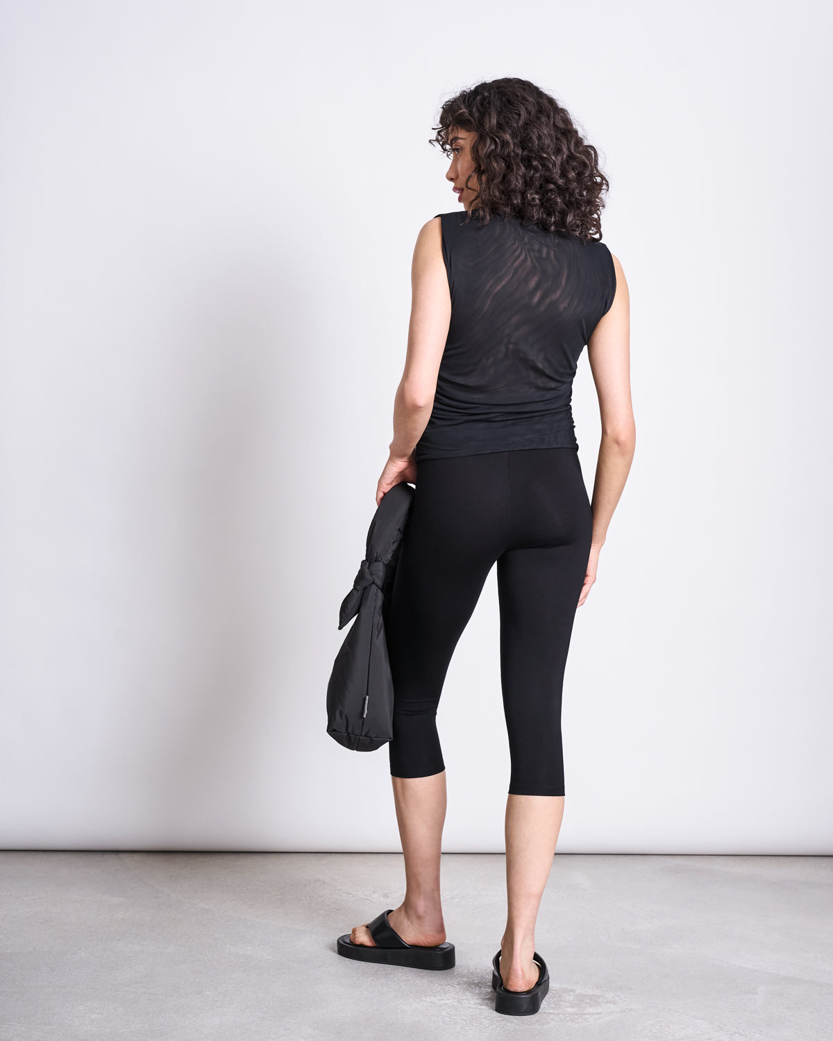 A person with curly hair, seen from behind, wears a sleeveless black top, jan-n-june CAPRI LEGGINGS BLACK made with TENCEL™ Lyocell, and black sandals. They hold a black bag and stand against a plain light background.