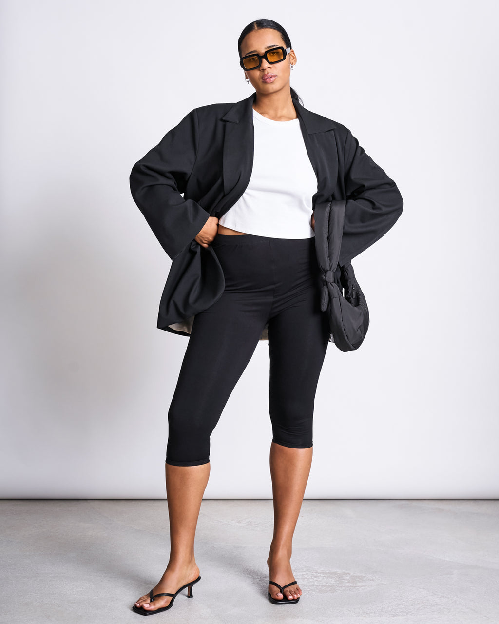 A woman models jan-n-june CAPRI LEGGINGS BLACK with sunglasses, a white crop top, black oversized blazer, black heeled sandals, and a black bag, standing confidently against a plain white background.