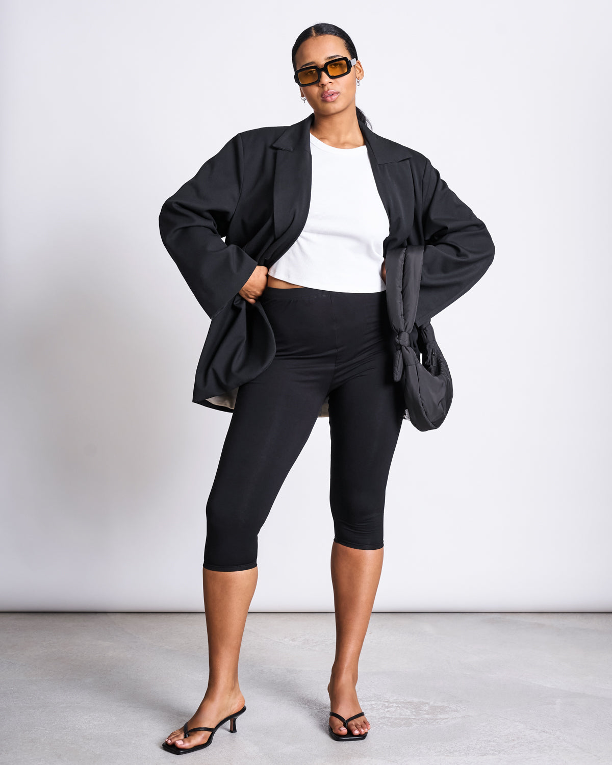 A woman models jan-n-june CAPRI LEGGINGS BLACK with sunglasses, a white crop top, black oversized blazer, black heeled sandals, and a black bag, standing confidently against a plain white background.