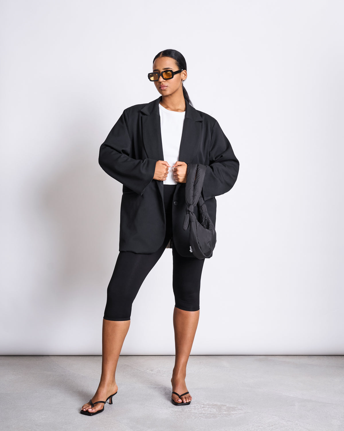 A woman stands confidently against a white background, wearing an oversized black blazer, white shirt, jan-n-june CAPRI LEGGINGS BLACK, heeled sandals, bold sunglasses, and holding a black bag.