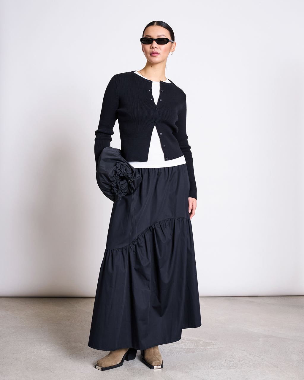 A woman in jan-n-june’s black CREWNECK CARDIGAN GLIMT (GOTS), sunglasses, a white top, long black tiered skirt, tan square-toe boots, and holding a large ruffled black clutch stands against a plain wall.