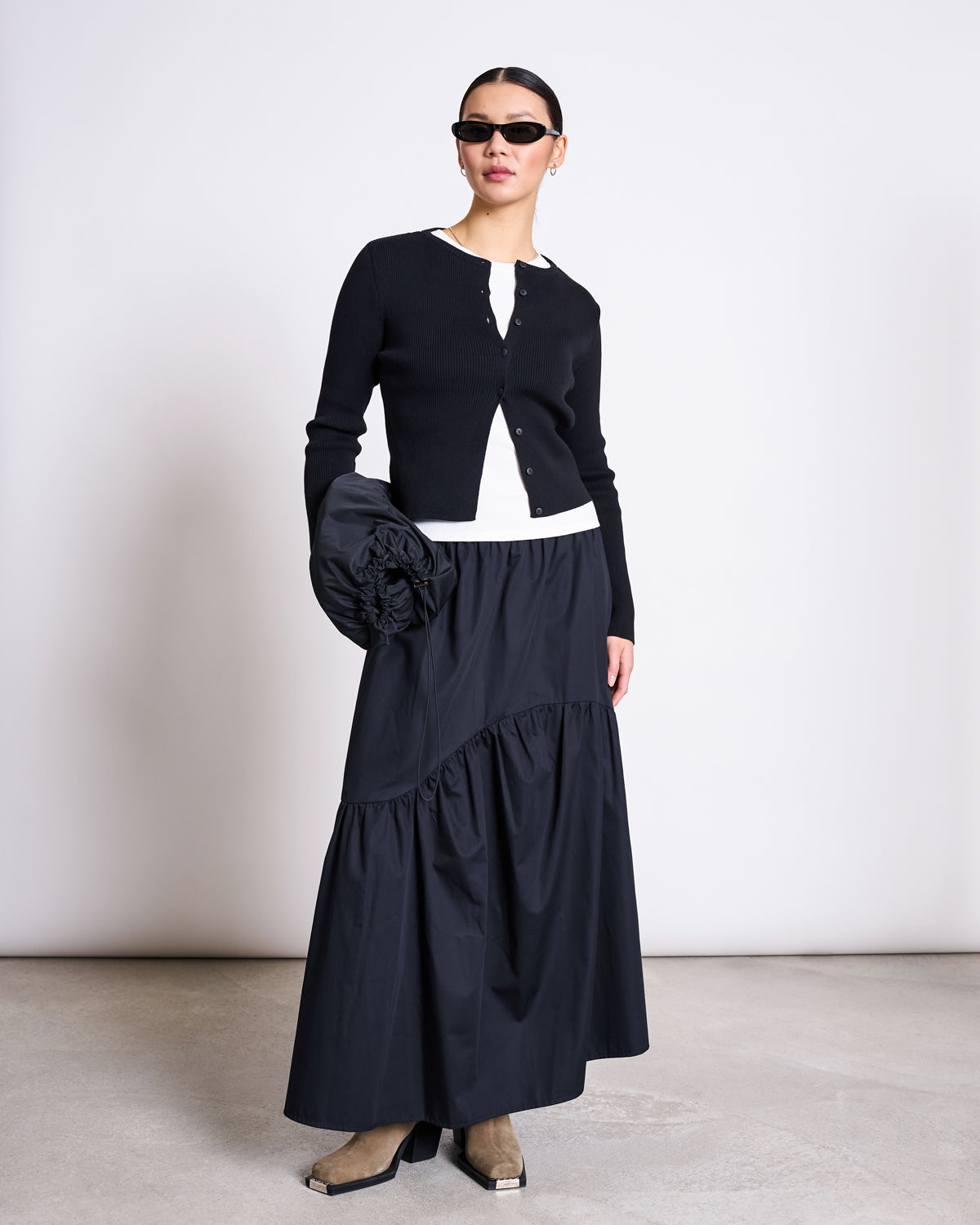 A woman in jan-n-june’s black CREWNECK CARDIGAN GLIMT (GOTS), sunglasses, a white top, long black tiered skirt, tan square-toe boots, and holding a large ruffled black clutch stands against a plain wall.