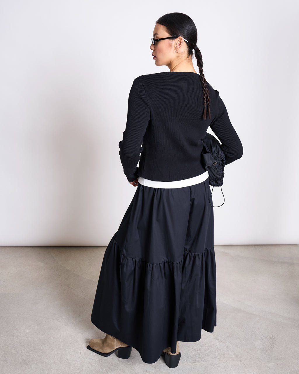 A woman with braided hair, seen from behind, wears the jan-n-june CREWNECK CARDIGAN GLIMT BLACK GOTS, a tiered black skirt, tan boots, sunglasses, and holds a black bag against a plain light background.