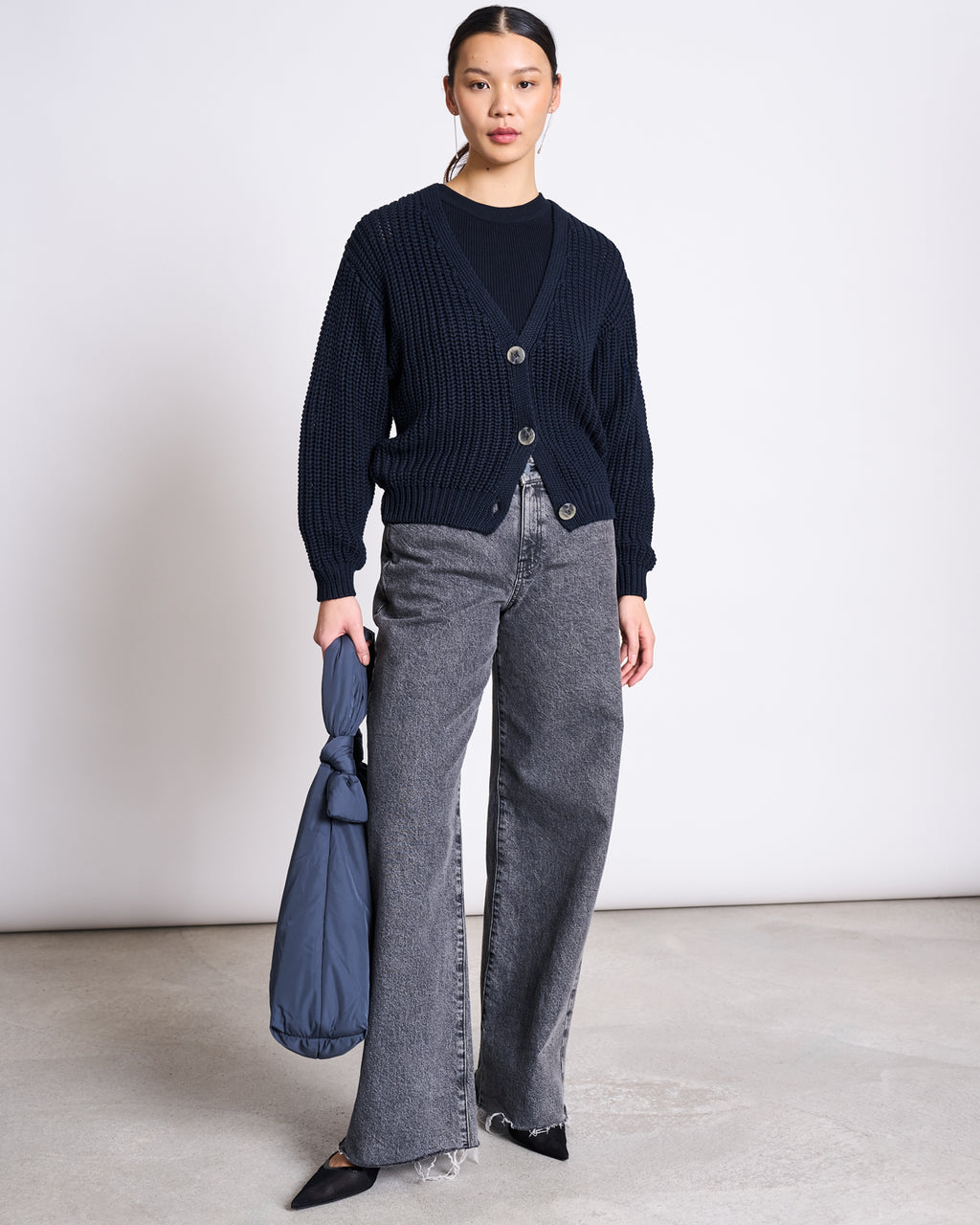 A woman stands indoors against a plain background, wearing the jan-n-june ORGANIC COTTON TANK TOP PORTO LONG NIGHTFALL GOTS under a black sweater, with gray wide-leg jeans, black pointed shoes, and holding a folded blue bag in her left hand.