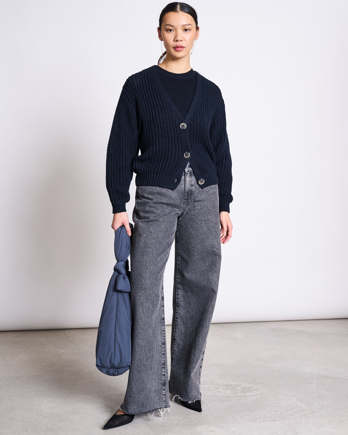 A woman stands indoors against a plain background, wearing the jan-n-june ORGANIC COTTON TANK TOP PORTO LONG NIGHTFALL GOTS under a black sweater, with gray wide-leg jeans, black pointed shoes, and holding a folded blue bag in her left hand.