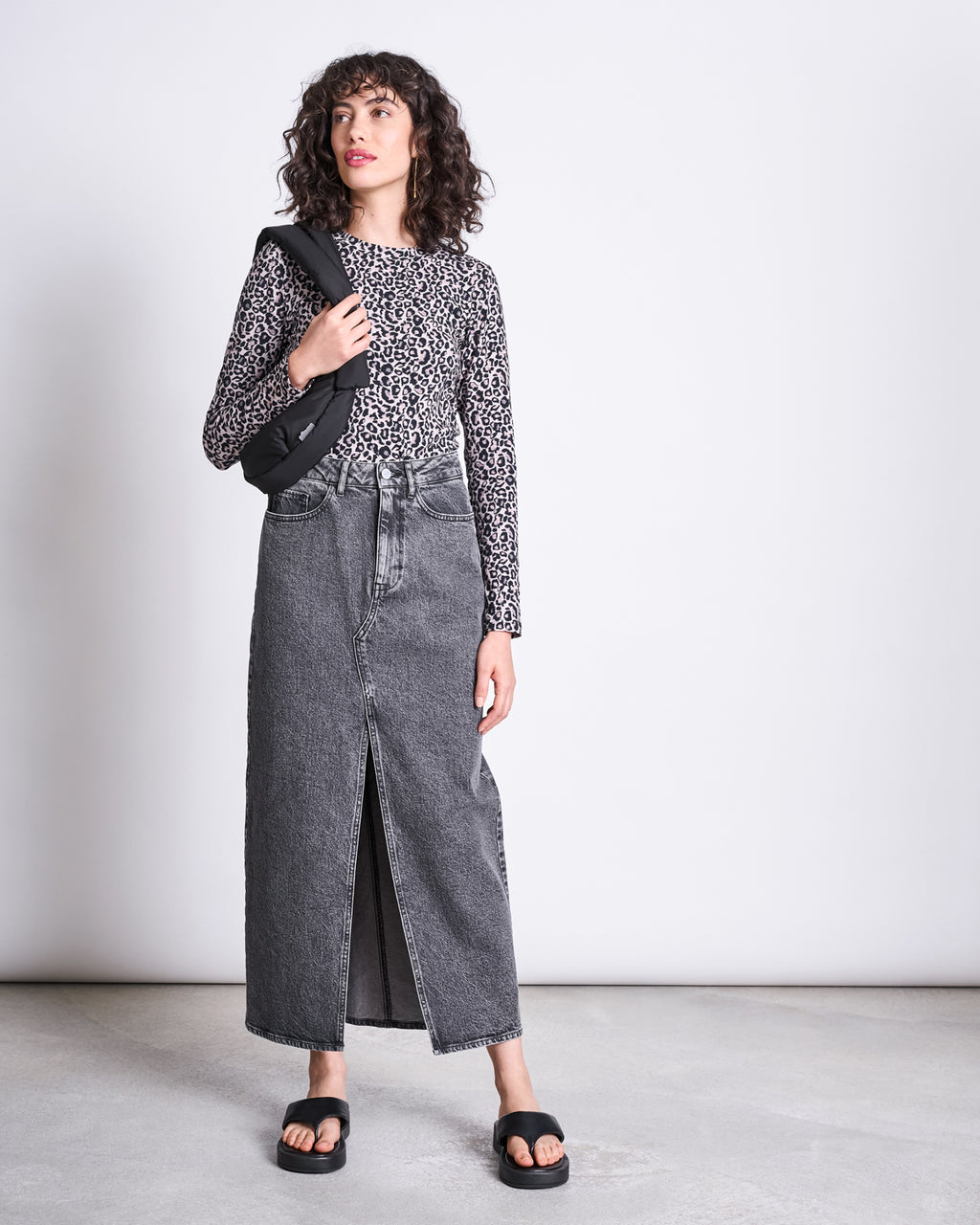 A person stands against a plain background wearing the jan-n-june LONGSLEEVE BARID LEO GOTS as a statement piece, styled with a high-waisted gray denim maxi skirt with a front slit, black sandals, and a black crossbody bag.
