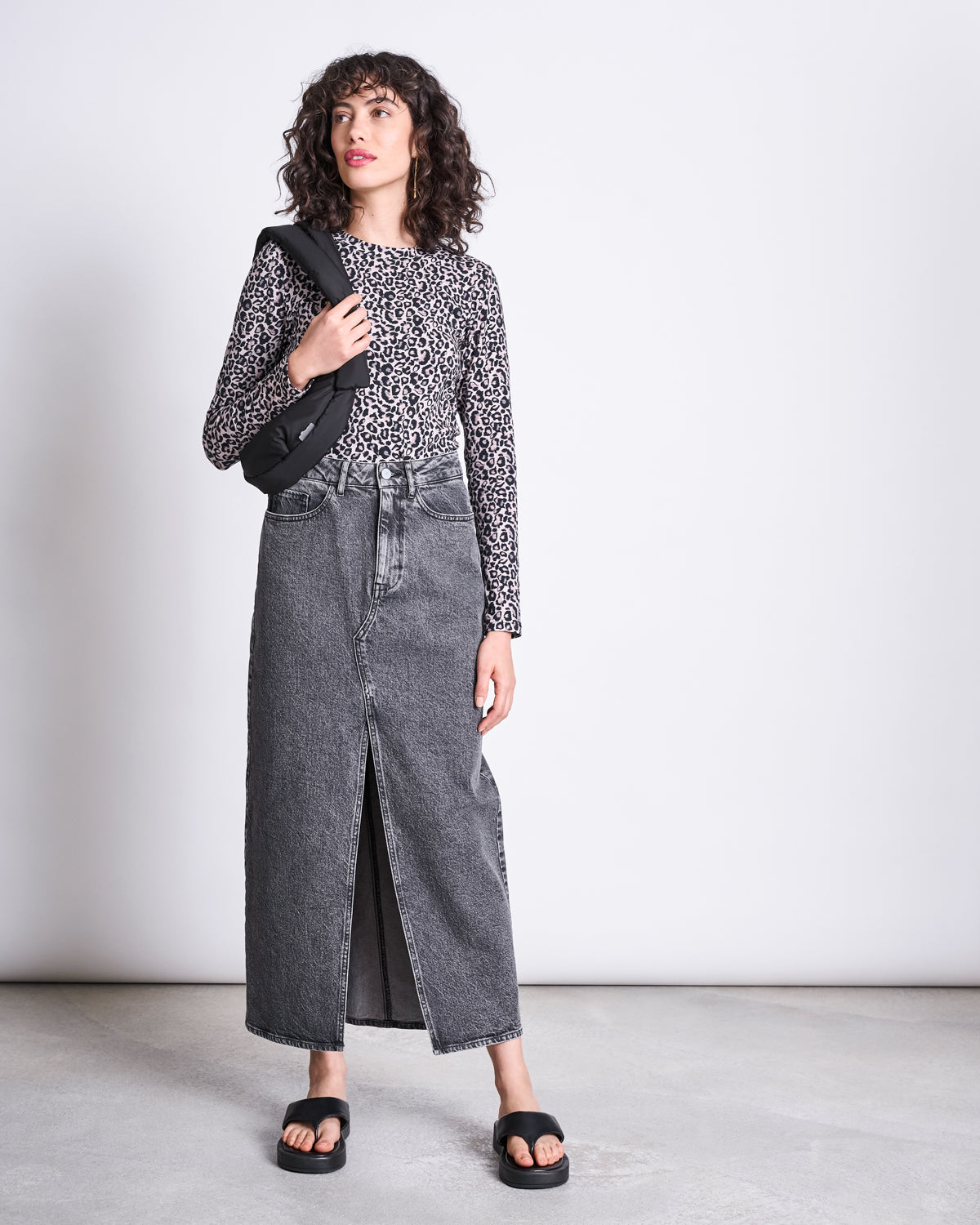 A person stands against a plain background wearing the jan-n-june LONGSLEEVE BARID LEO GOTS as a statement piece, styled with a high-waisted gray denim maxi skirt with a front slit, black sandals, and a black crossbody bag.