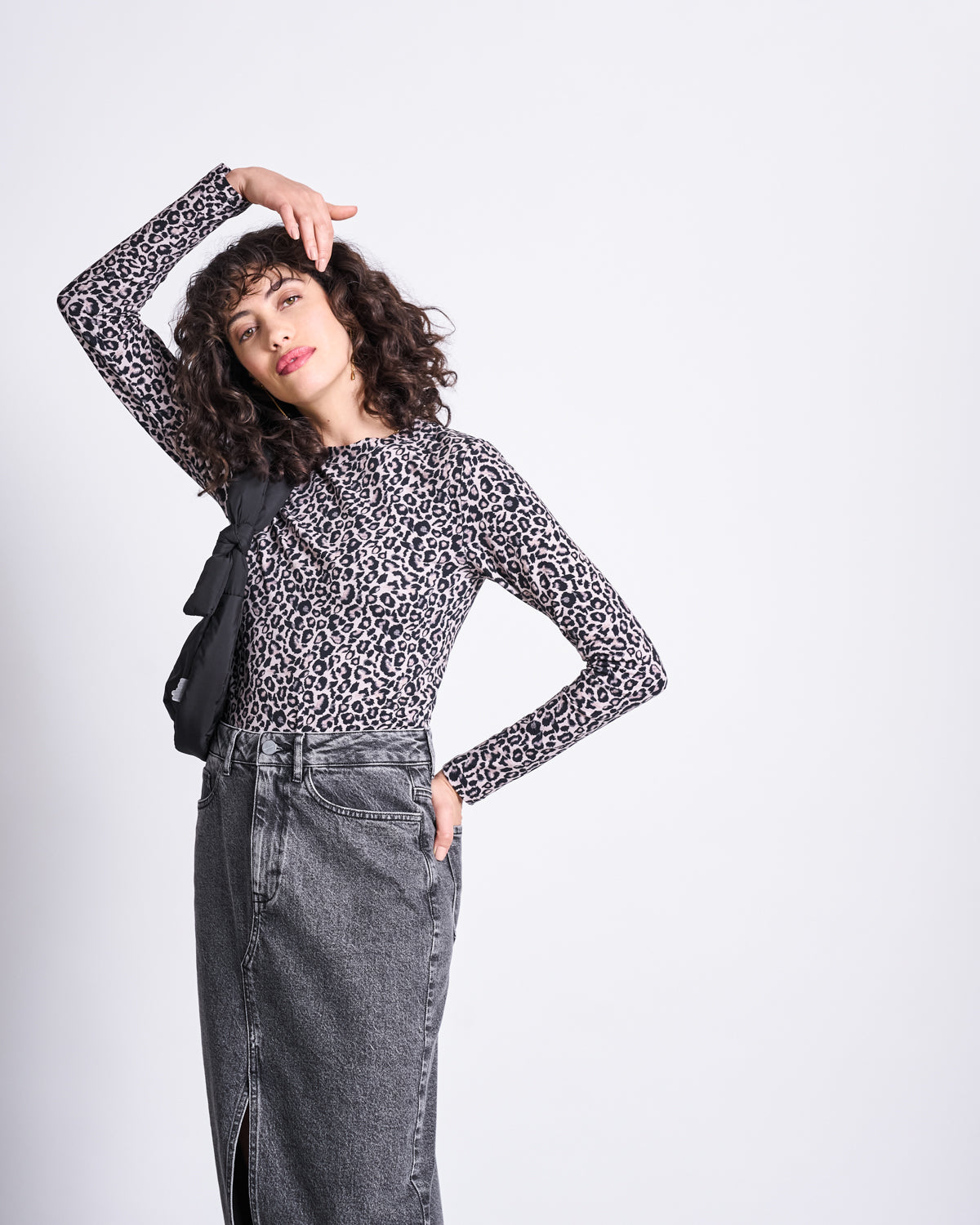 A woman with curly hair poses against a plain background, wearing the jan-n-june LONGSLEEVE BARID LEO GOTS as a statement piece, paired with a gray denim skirt with a front slit and a black bag. One arm is raised, the other rests in her pocket.