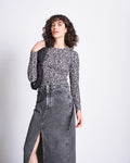 A woman with curly hair wears the jan-n-june LONGSLEEVE BARID LEO GOTS and a high-waisted gray denim skirt with a front slit, holding a black bag over her shoulder against a plain light background.