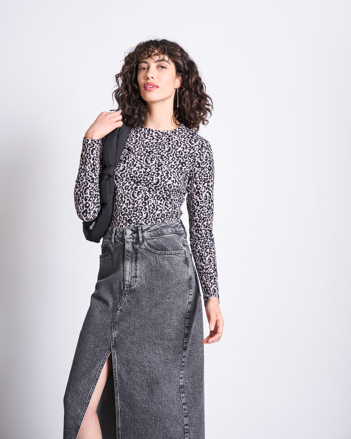 A woman with curly hair wears the jan-n-june LONGSLEEVE BARID LEO GOTS and a high-waisted gray denim skirt with a front slit, holding a black bag over her shoulder against a plain light background.