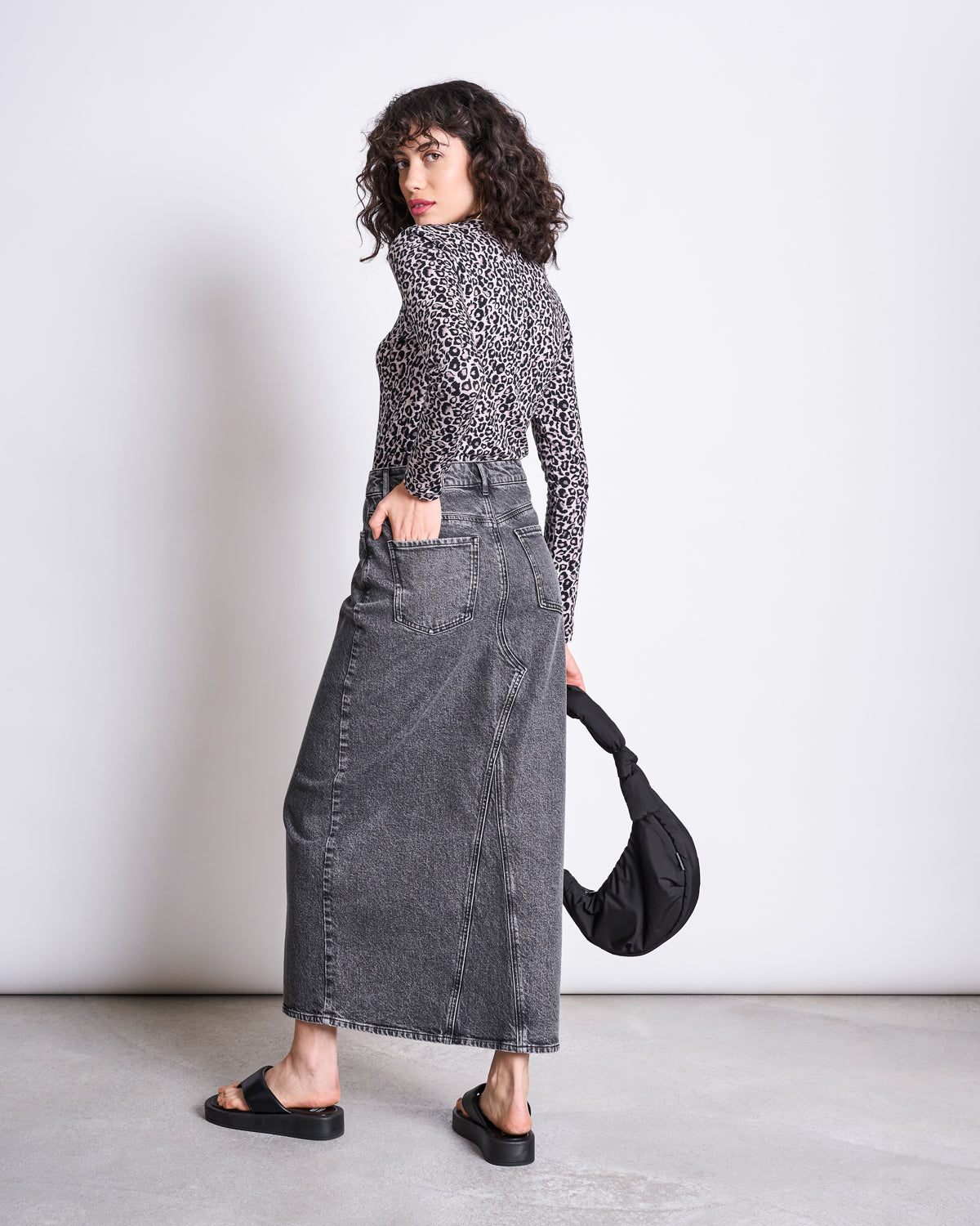 A woman with curly hair wears the jan-n-june LONGSLEEVE BARID LEO GOTS and a high-waisted gray denim midi skirt, paired with black sandals and holding a black bag, standing against a plain background.