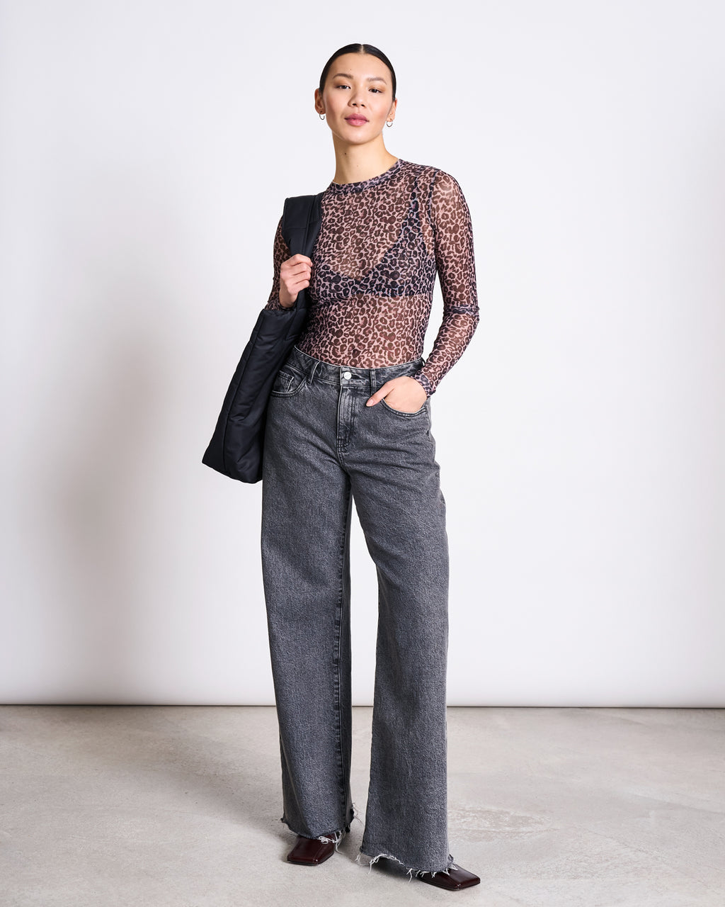 A woman poses against a plain background in the jan-n-june MESH LONGSLEEVE DORA LEO PRINT top, high-waisted wide-leg gray jeans with frayed hems, dark shoes, and holds a black jacket over her shoulder.