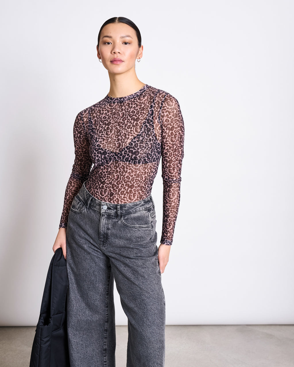 A woman poses confidently against a plain background in the jan-n-june MESH LONGSLEEVE DORA LEO PRINT paired with high-waisted gray jeans, holding a black jacket in one hand.