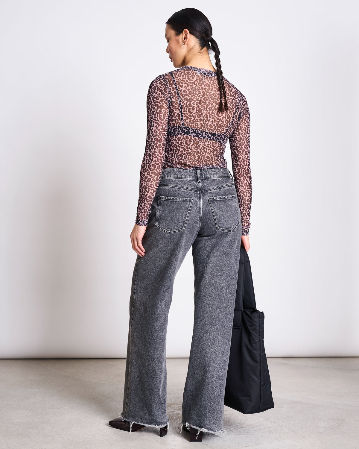 A person with braided hair faces away, wearing the jan-n-june MESH LONGSLEEVE DORA LEO PRINT top and high-waisted, wide-leg gray jeans with frayed hems, holding a black bag in their left hand.