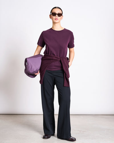 A person in sunglasses wears the jan-n-june BRUSHED T-SHIRT BOY CHERRY LACQUER GOTS, with a matching sweater at the waist, black pants, and dark shoes, holding a folded purple jacket against a plain light background.