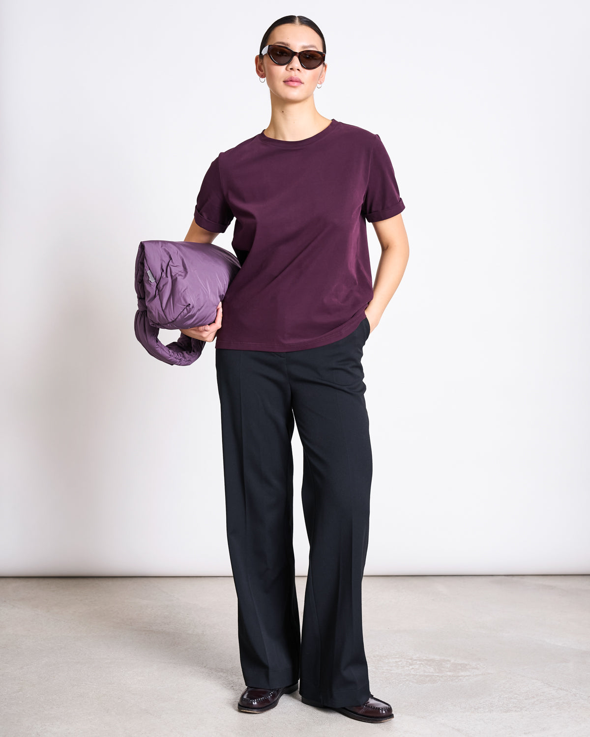 A person in sunglasses and black trousers stands against a plain background, wearing the jan-n-june BRUSHED T-SHIRT BOY CHERRY LACQUER GOTS in purple and holding a rolled-up purple jacket or coat.