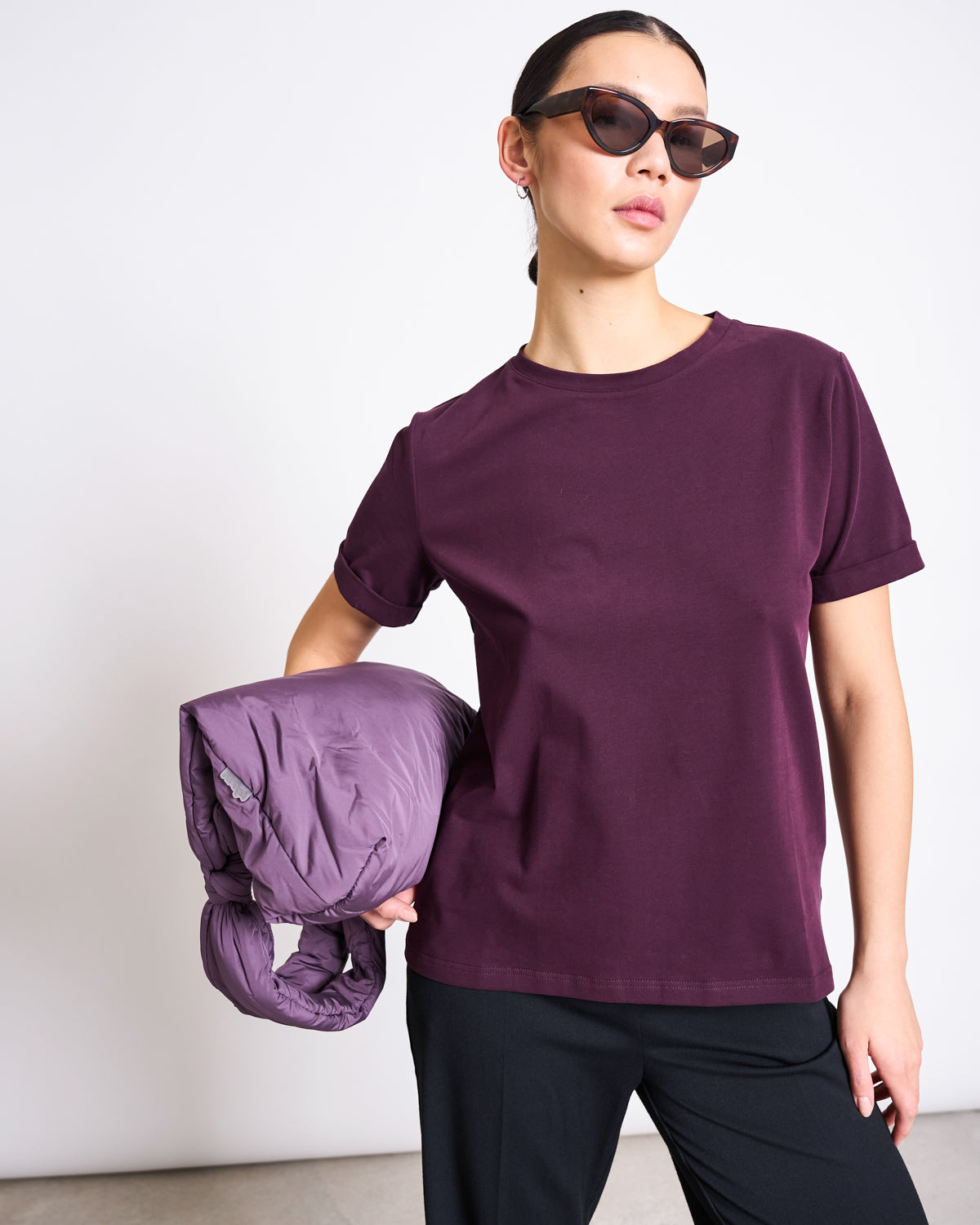 A woman in dark sunglasses and black pants holds a purple puffer jacket under her arm, wearing the jan-n-june BRUSHED T-SHIRT BOY CHERRY LACQUER GOTS, standing against a plain white background.