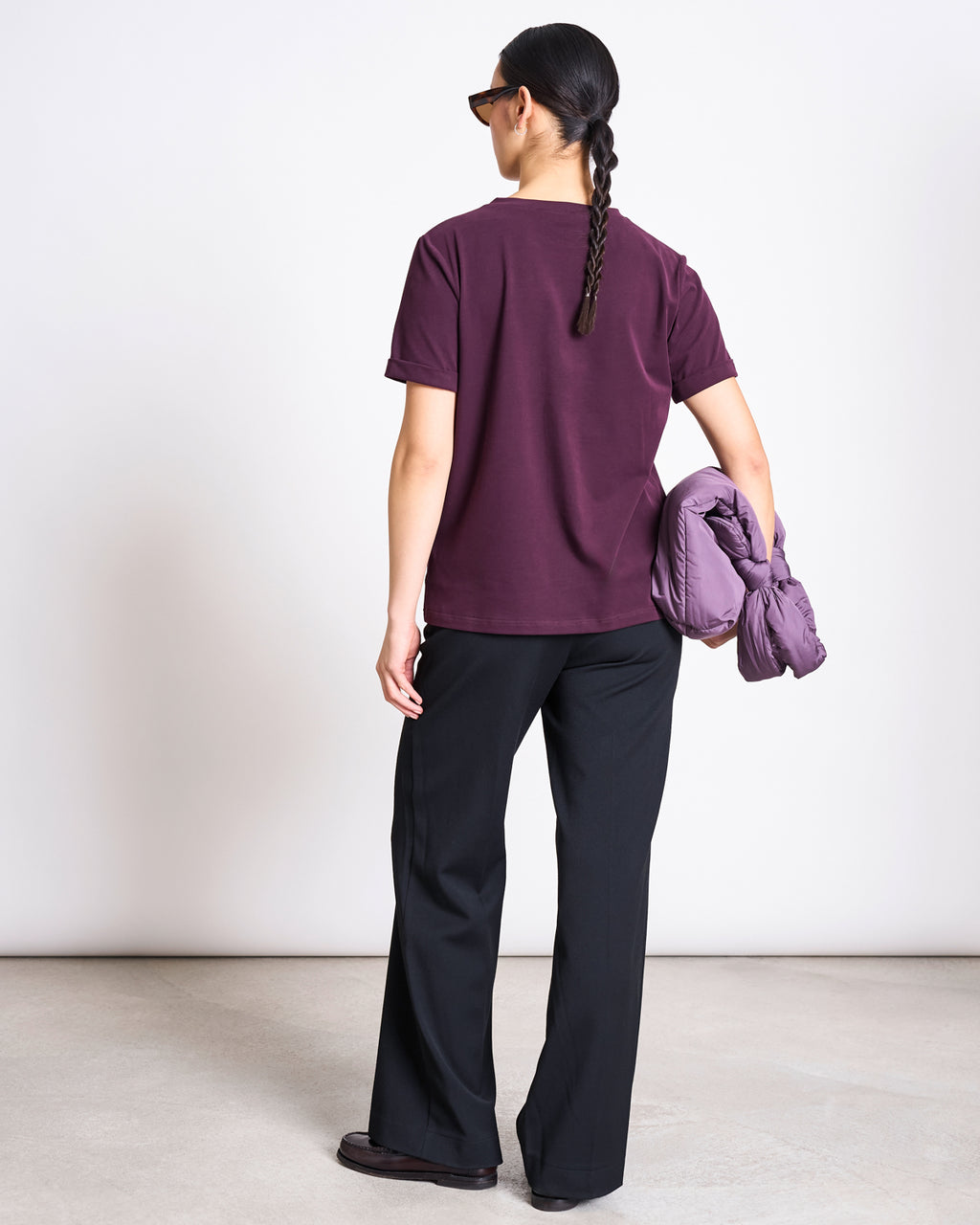 A person with a long braid, in sunglasses, wears jan-n-junes BRUSHED T-SHIRT BOY CHERRY LACQUER GOTS and black pants, holding a matching maroon jacket under their arm against a plain light background.