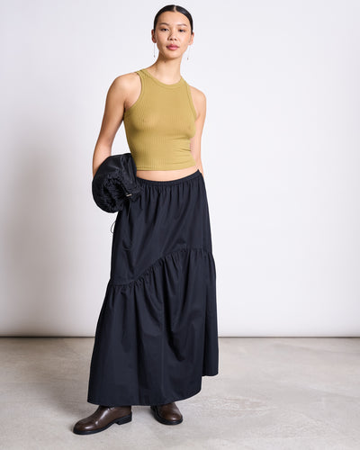 A woman stands confidently against a plain background in the jan-n-june TIERED MAXI SKIRT ALCA BLACK, styled with a chic sleeveless olive green crop top, brown boots, and a black jacket. Her hair is tied back as she looks boldly at the camera.
