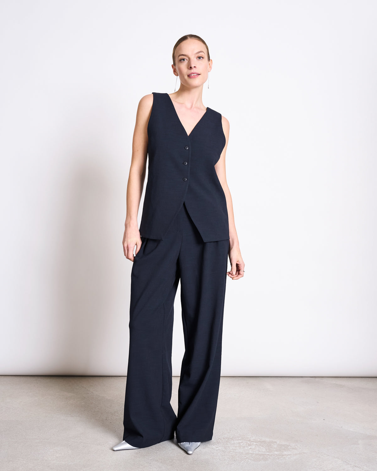 A woman with light hair pulled back stands against a white wall wearing jan-n-june’s sleeveless dark navy V-neck vest and PLEATED WIDE PANTS NIVA NIGHTFALL, paired with pointed silver shoes.