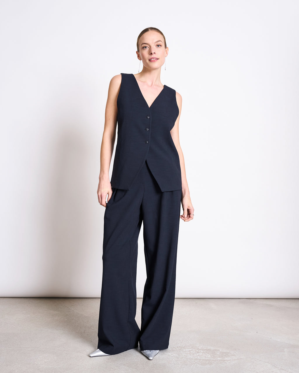 A woman stands in a minimalist room wearing the jan-n-june SUIT VEST IMATA NIGHTFALL with a deep V-neck and three buttons, paired with matching wide-leg trousers and pointed silver heels for an elegant formal look.