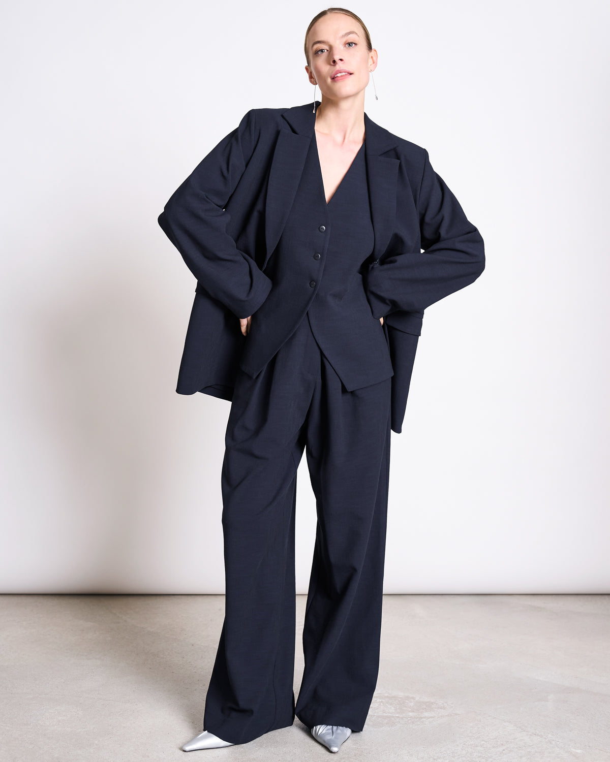 A person stands confidently against a plain background, wearing jan-n-junes PLEATED WIDE PANTS NIVA NIGHTFALL made from EcoVero™, paired with a buttoned vest, blazer, and pointed silver shoes.