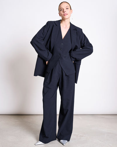 A person stands confidently against a plain background, wearing jan-n-junes PLEATED WIDE PANTS NIVA NIGHTFALL made from EcoVero™, paired with a buttoned vest, blazer, and pointed silver shoes.