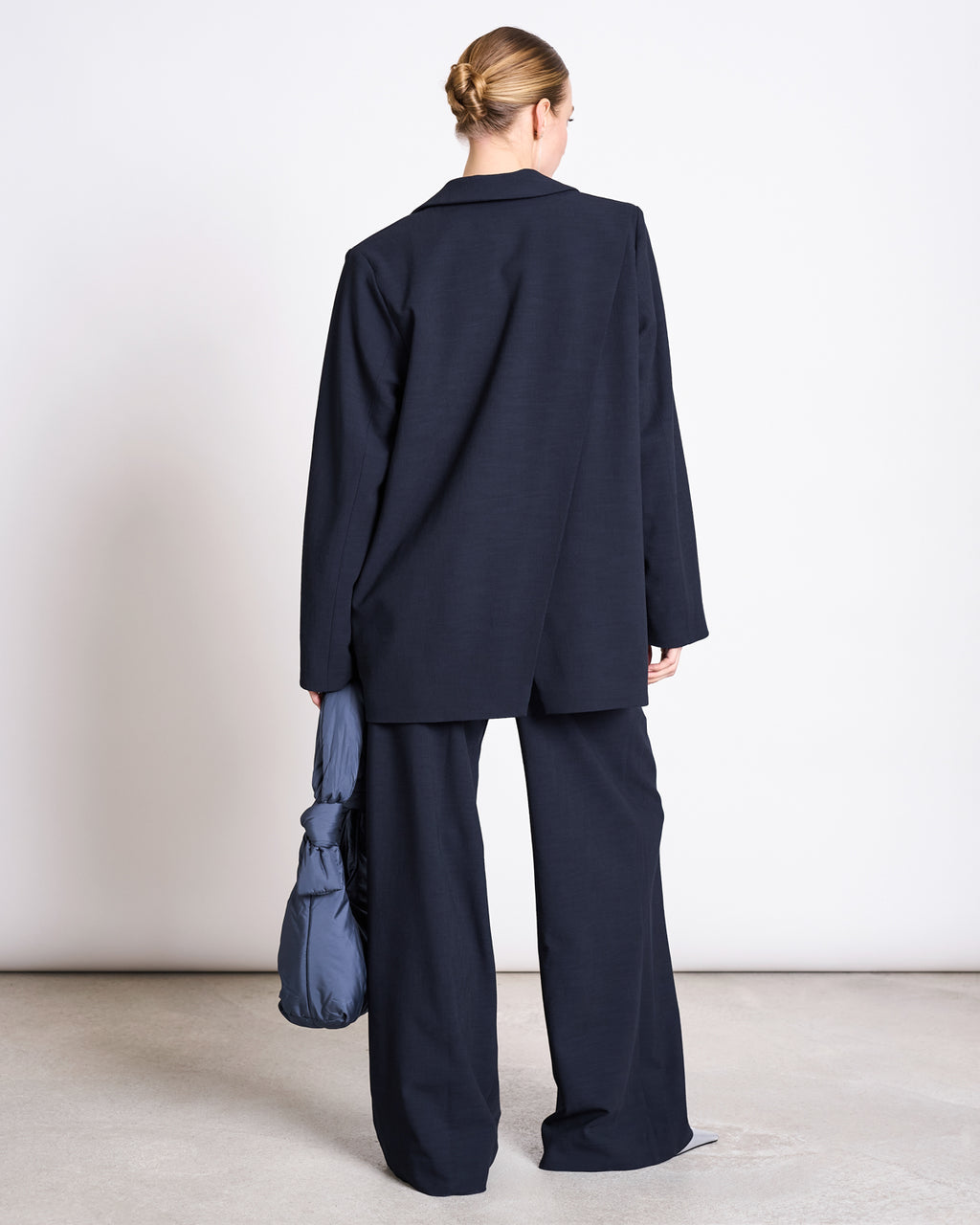 A person with blonde hair in a neat bun stands with their back to the camera, wearing the jan-n-june SLITTED BACK BLAZER JELS NIGHTFALL and holding a puffy blue bag. The background is plain white.