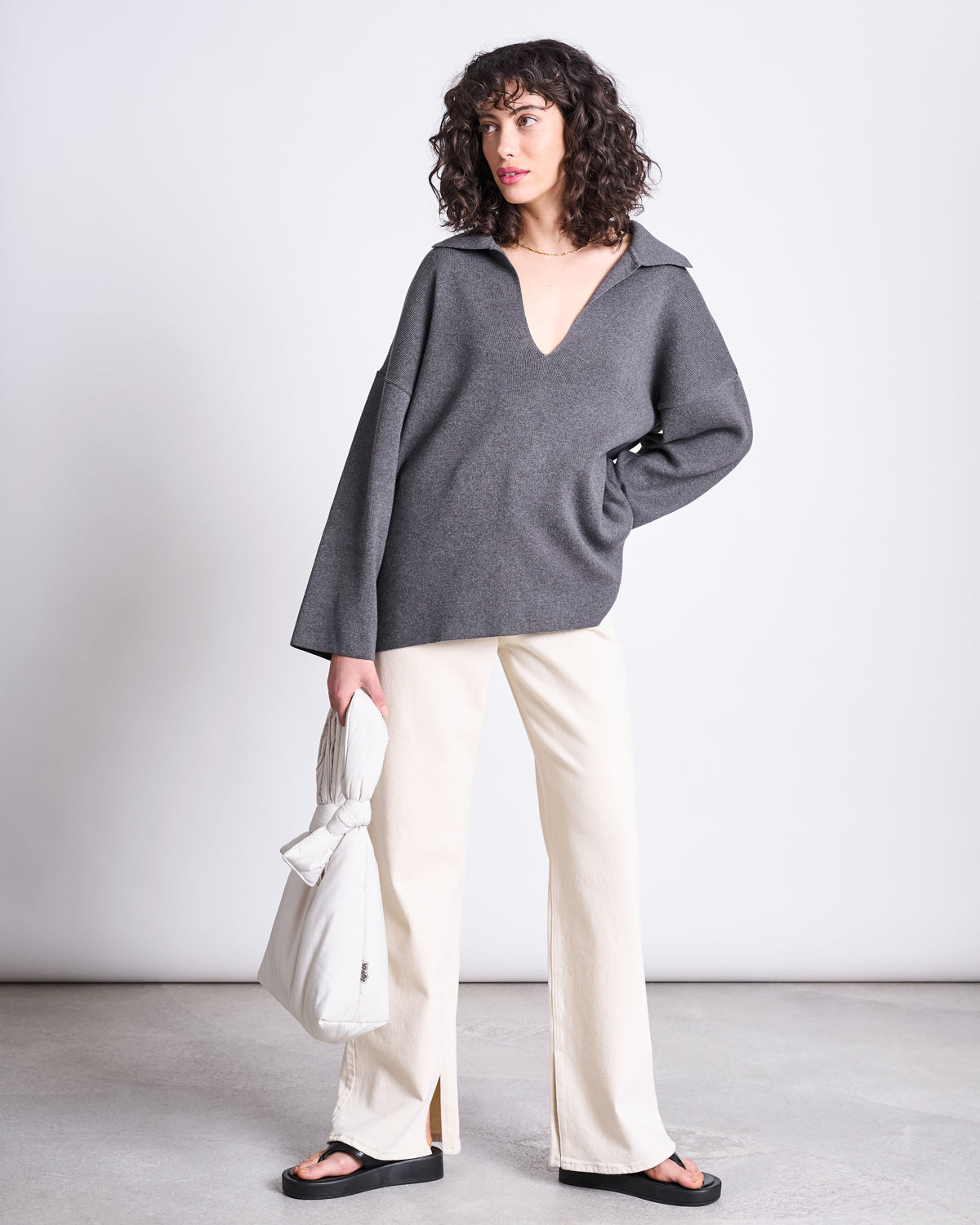 A woman with curly hair wears a loose gray V-neck sweater, off-white wide-leg pants, and black sandals. She holds the jan-n-june BAG BIWA PUFFY MINI ICE in white and stands against a plain light background.