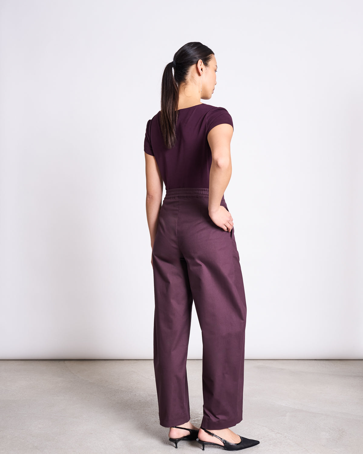 A woman faces away from the camera in a jan-n-june BRUSHED BOAT NECK T-SHIRT OTTO CHERRY LACQUER GOTS, paired with matching high-waisted pants and black heels. Her hair is tied back, set against a plain light background.