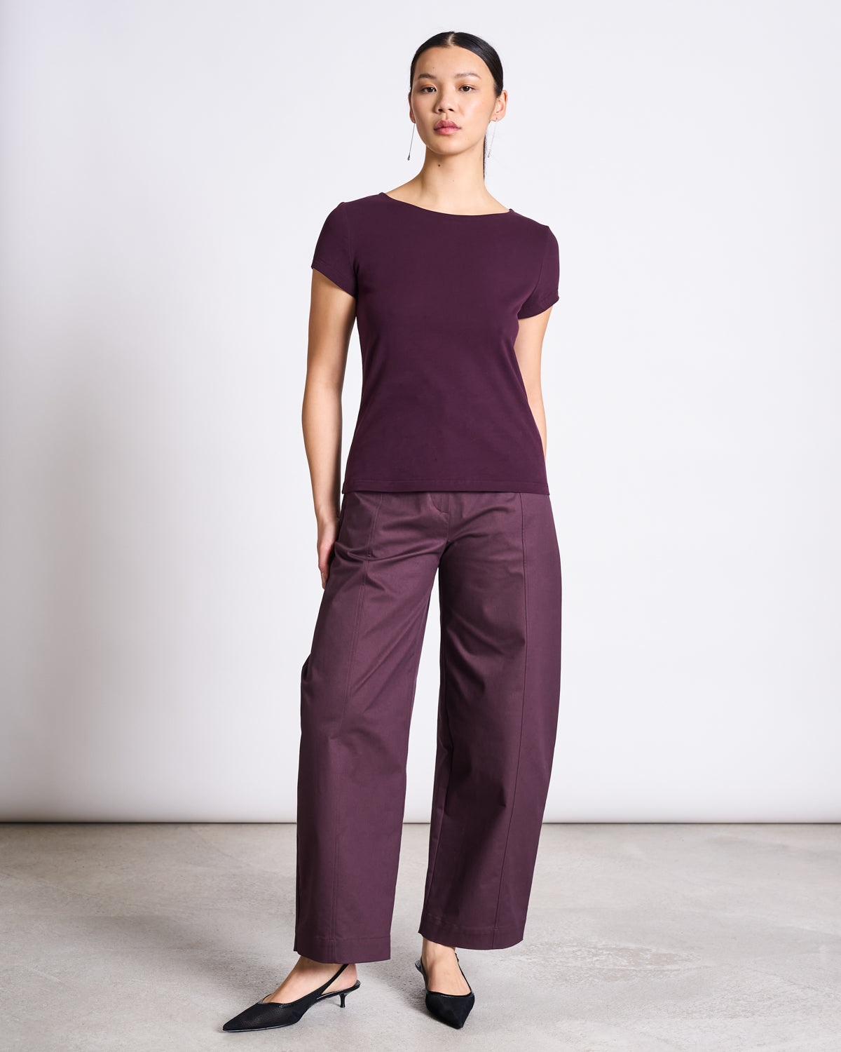 A woman stands against a plain background wearing the jan-n-june BRUSHED BOAT NECK T-SHIRT OTTO CHERRY LACQUER GOTS—short-sleeved, super-soft, with a classic boat neckline—paired with matching wide-leg trousers and black pointed-toe heels.
