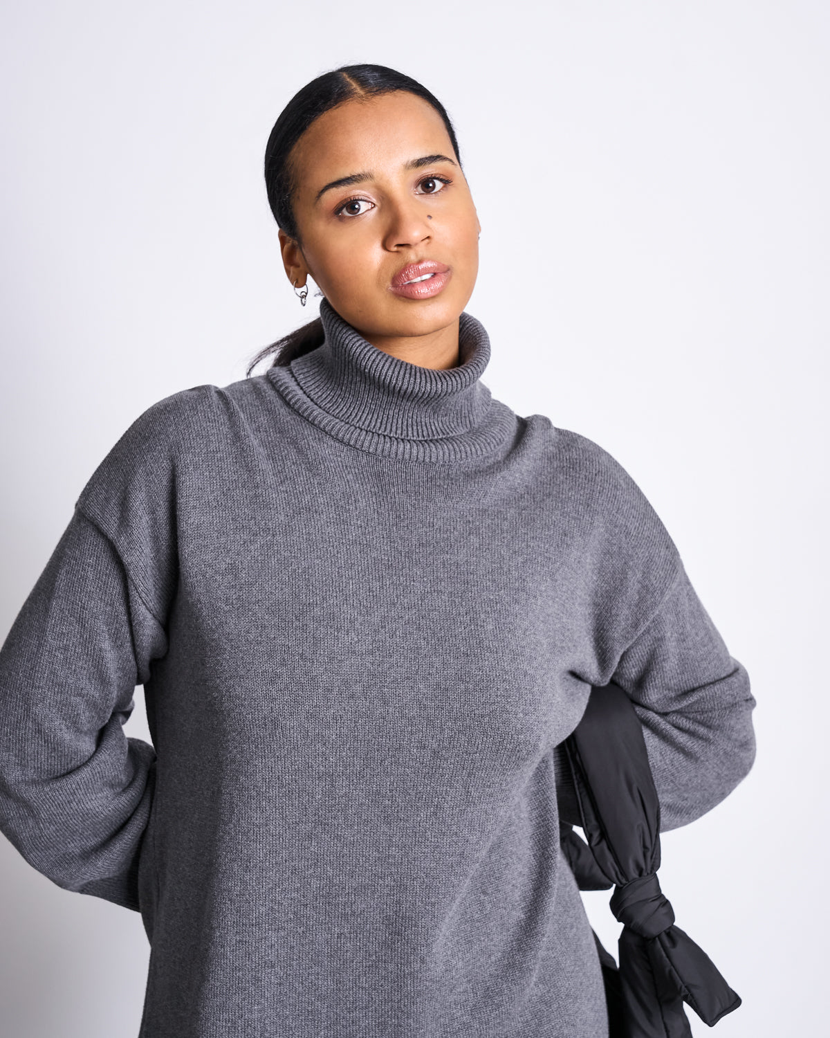 A woman with dark hair tied back wears the jan-n-june MIDI KNIT DRESS INARI DARK GREY GOTS and small hoop earrings, standing against a plain light background with a neutral expression, holding a black item draped over her left arm.