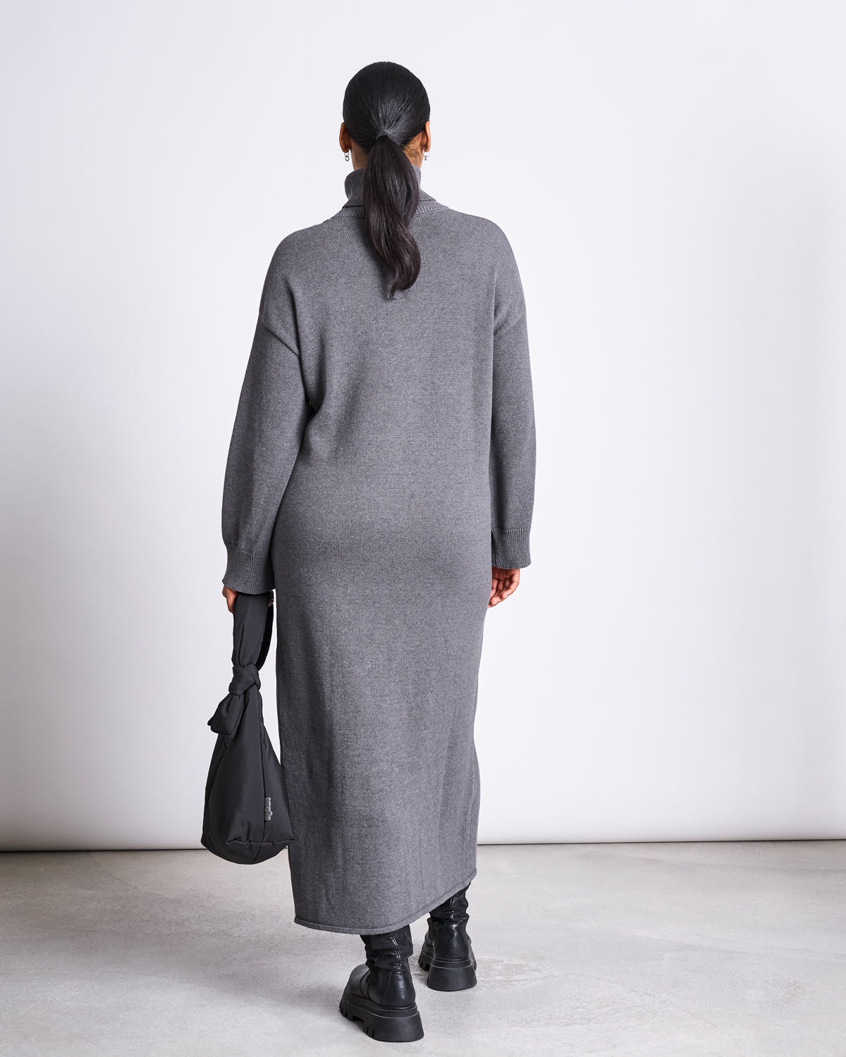 A person with tied-back hair stands against a plain white background, wearing the jan-n-june MIDI KNIT DRESS INARI DARK GREY GOTS, paired with black boots and holding a black bag.