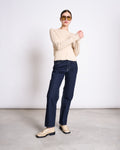A woman poses with arms raised and crossed at the wrists, wearing jan-n-june’s GOTS Certified CREWNECK CARDIGAN GLIMT BEIGE, dark blue jeans, beige loafers, and large yellow-tinted sunglasses against a plain background.
