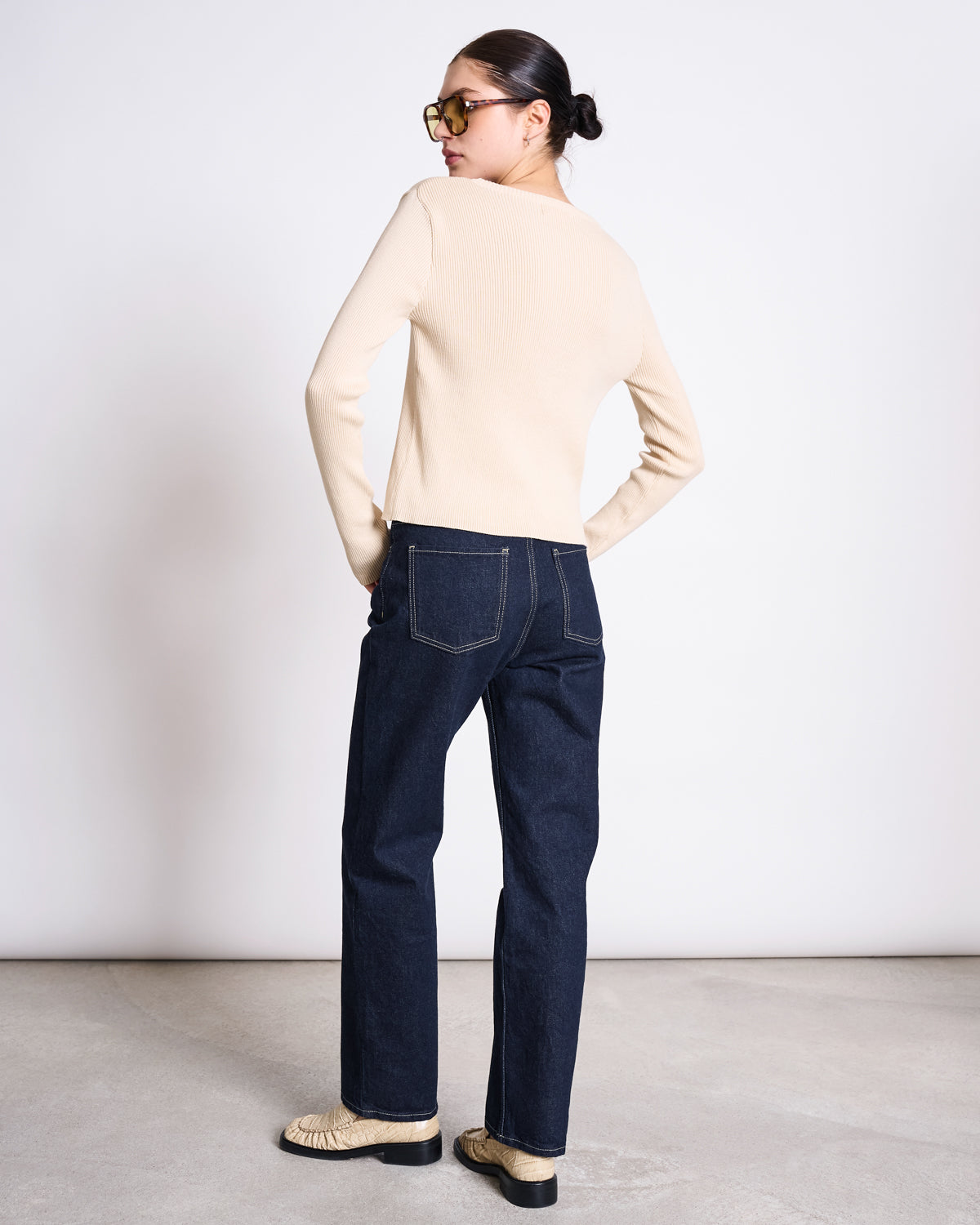 A person with dark hair in a low bun, sunglasses, and woven shoes stands facing away with hands in back pockets, wearing jan-n-june's CREWNECK CARDIGAN GLIMT BEIGE GOTS and dark blue jeans with visible stitching.