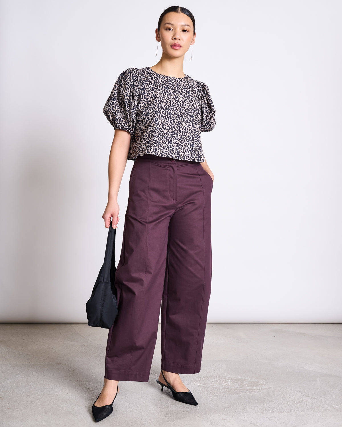 An eco-conscious fashionista stands against a plain background, wearing the jan-n-june CROPPED ORGANIC COTTON BLOUSE OLMOS LEO PRINT, styled with high-waisted maroon wide-leg pants, black heeled shoes, and holding a black bag.