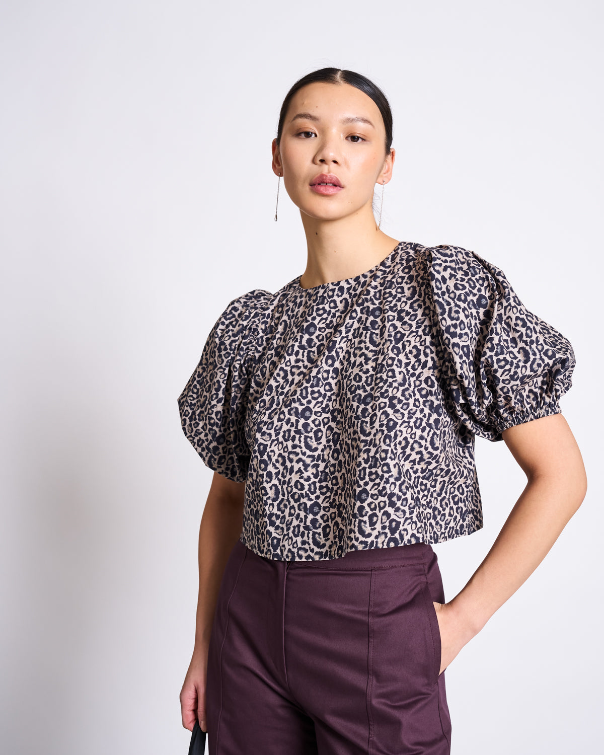 A style-savvy trendsetter rocks the jan-n-june CROPPED ORGANIC COTTON BLOUSE OLMOS LEO PRINT with puff sleeves and high-waisted dark purple trousers, posing confidently against a plain white backdrop.